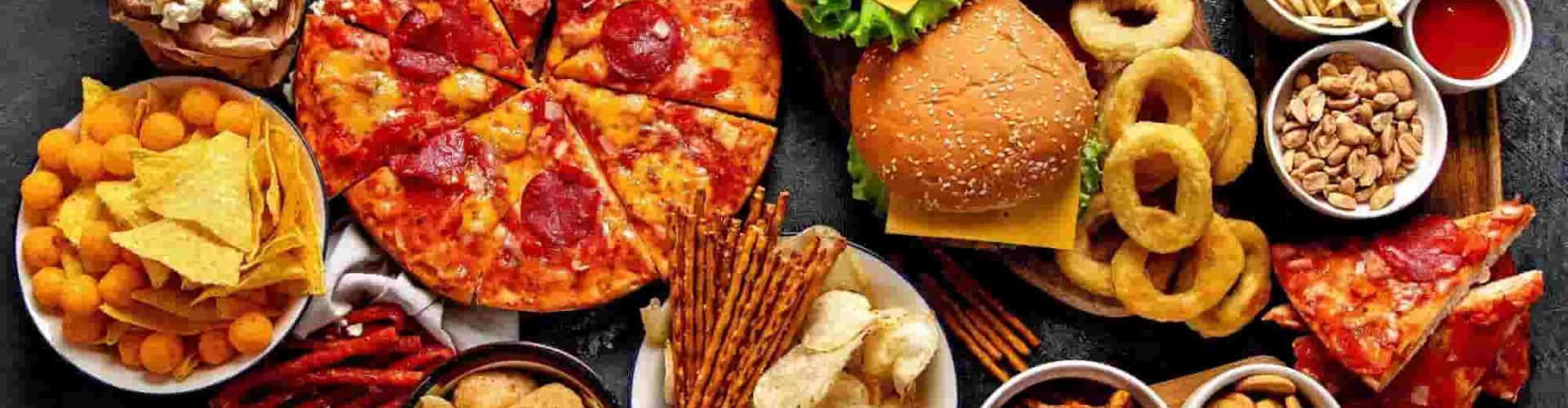 Delicious fast food platter featuring pepperoni pizza, cheeseburgers, French fries, onion rings, nachos, popcorn, potato chips, pretzels, and assorted snacks on a dark table – perfect for a fast food restaurant near me
